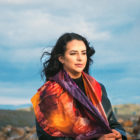 Young woman with black hair wearing a silk scarf. Sitting on rocks with a backdrop of sky and cloud