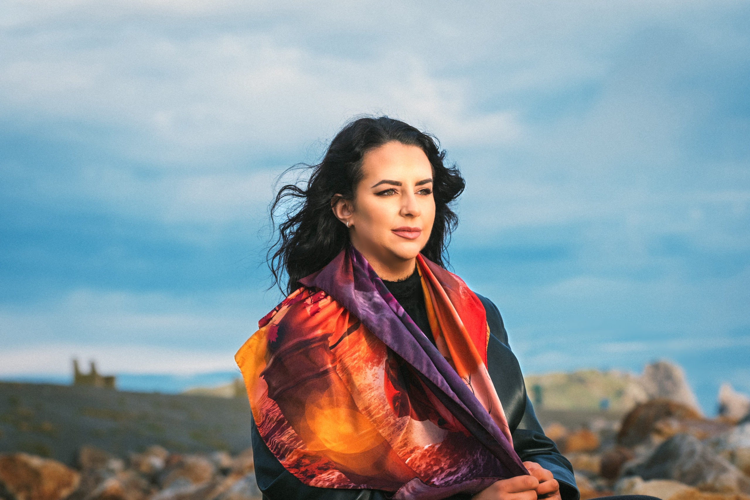 Young woman with black hair wearing a silk scarf. Sitting on rocks with a backdrop of sky and cloud