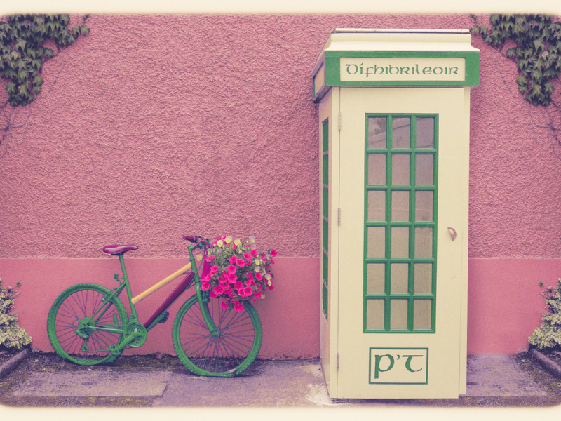 An old Irish telephone box set next to a pink wall and a colourful bike.