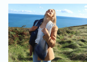Young woman with head thrown back laughing. Holding a meditation scarf around her neck. Green field and blue sea in the background.
